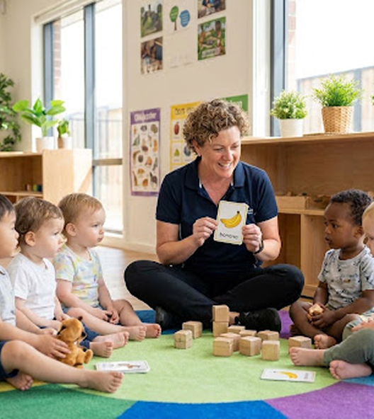 A professional and warm high-quality photograph of a friendly care assistant or early years educator sitting on a colorful rug with a group of young children engaged in a playful learning activity. Children on a mat learning with a caring assistant